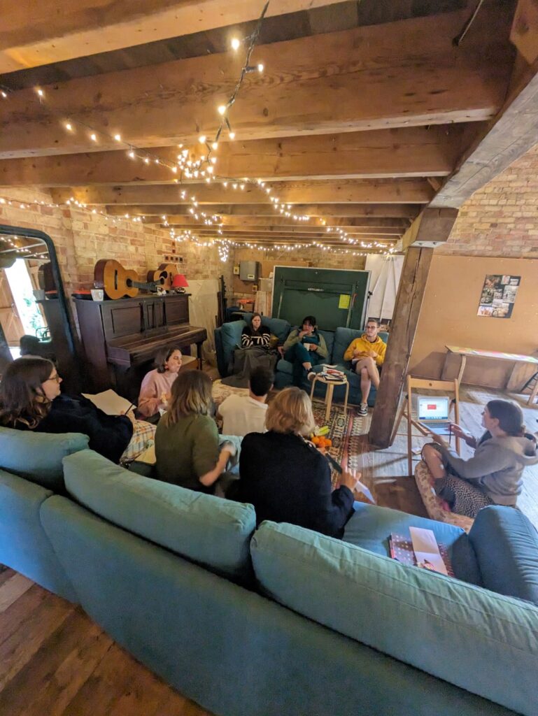 A group of people having a discussion. Part of the group is sat on a blue sofa facing away from the camera, and a small group is sat on a sofa opposite, facing the camera. Two people are sat on the floor, one at either end of the space between the sofas. The ceiling is low and there are fairy lights hanging from its wooden beams.