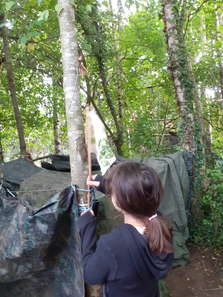 A child puts the finishing touch, a flag, on the corner of a shelter they have built in the forest. It is made of tarpaulins stretched between tree trunks.