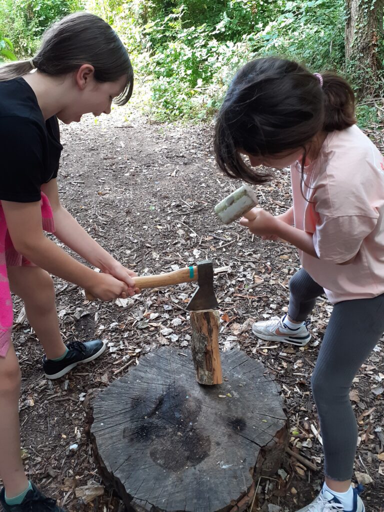 Two children are chopping a piece of wood. One has placed an axe on the end of the wood and the other is ready to swing a mallet on top of the axe.