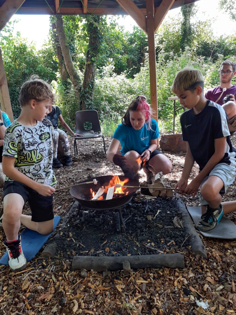 Two children and an adult kneel around a fire pit in a forest, under a tall wooden structure. They are toasting marshmallows.