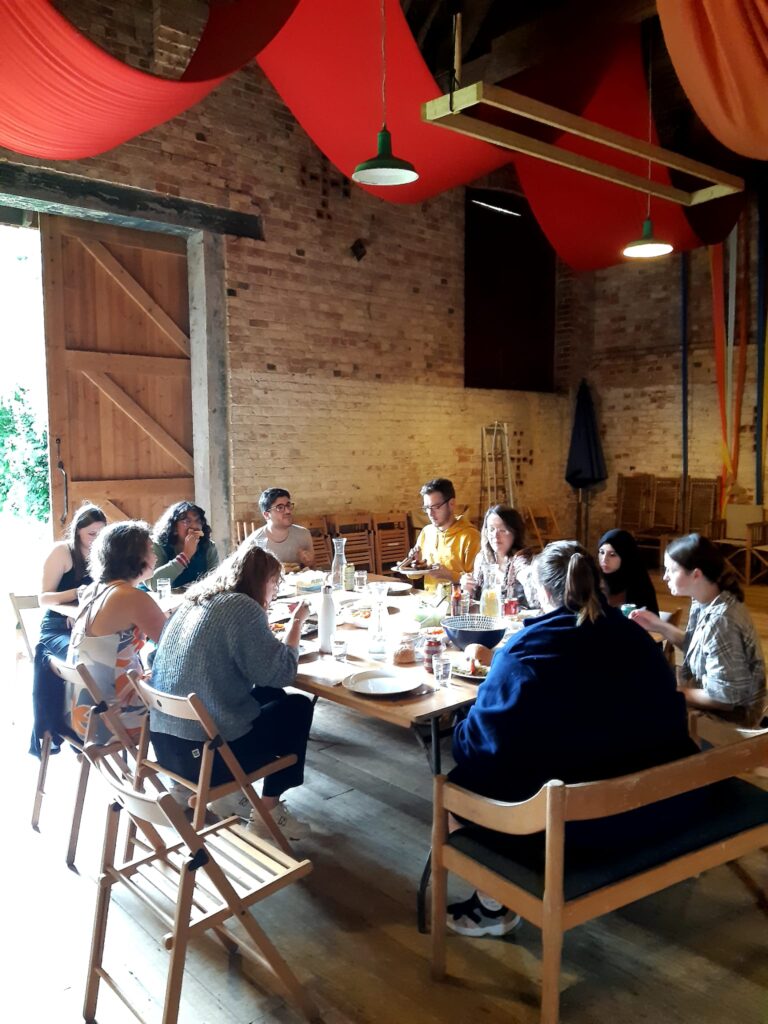 A group of people share a meal at a large wooden table. They are in a barn with brick walls and red drapes hanging from the ceiling.