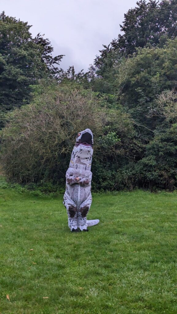Someone in a T-Rex costume stands in a grassy field in front of some trees and bushes.