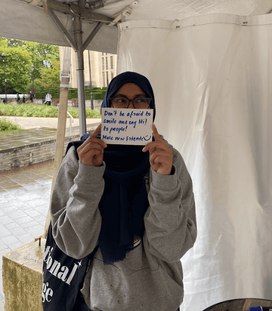 ID: a student holding a piece of paper in front of their face, their eyes are visible over the top. The paper reads 'Don't be afraid to say hi! to people. Make new friends'. There is a smiley face at the end of the writing.