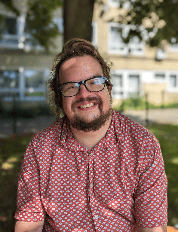 ID: Stephen is smiling at the camera. He is wearing glasses and has a bears. He is wearing a red patterned shirt.