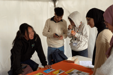 A group of students gather round an orange table, they are looking at printed materials on the table.