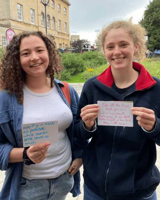 ID: two students are smiling at the camera. They are holding up pieces of paper. One reads 'enjoy first year as much as possible while marks don't count as much'; the other reads 'Get all the free food you can! Food is important. Don't worry if you don't make friends at the start, they will come!'