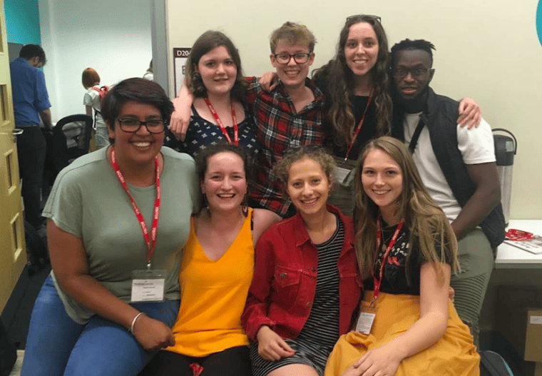 Sim and other staff members during her first year as a Projects Officer. She is wearing a tshirt and jeans, and is smiling at the camera. Seven other staff members are crowded together with Sim, also smiling.