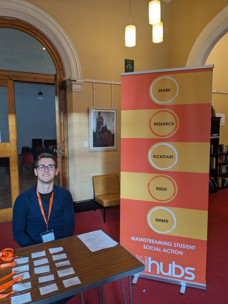 ID: Bristol Hub staff member Will sits at the venue's welcome desk. Next to him is a pull-up banner showing Student Hubs' model of delivery and reading Spark; Research; Kickstart; Build; Embed.