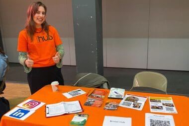 ID: a volunteer wearing an orange Bristol Hub t-shirt stands behind an orange table which is full of information about Bristol Hub activities.