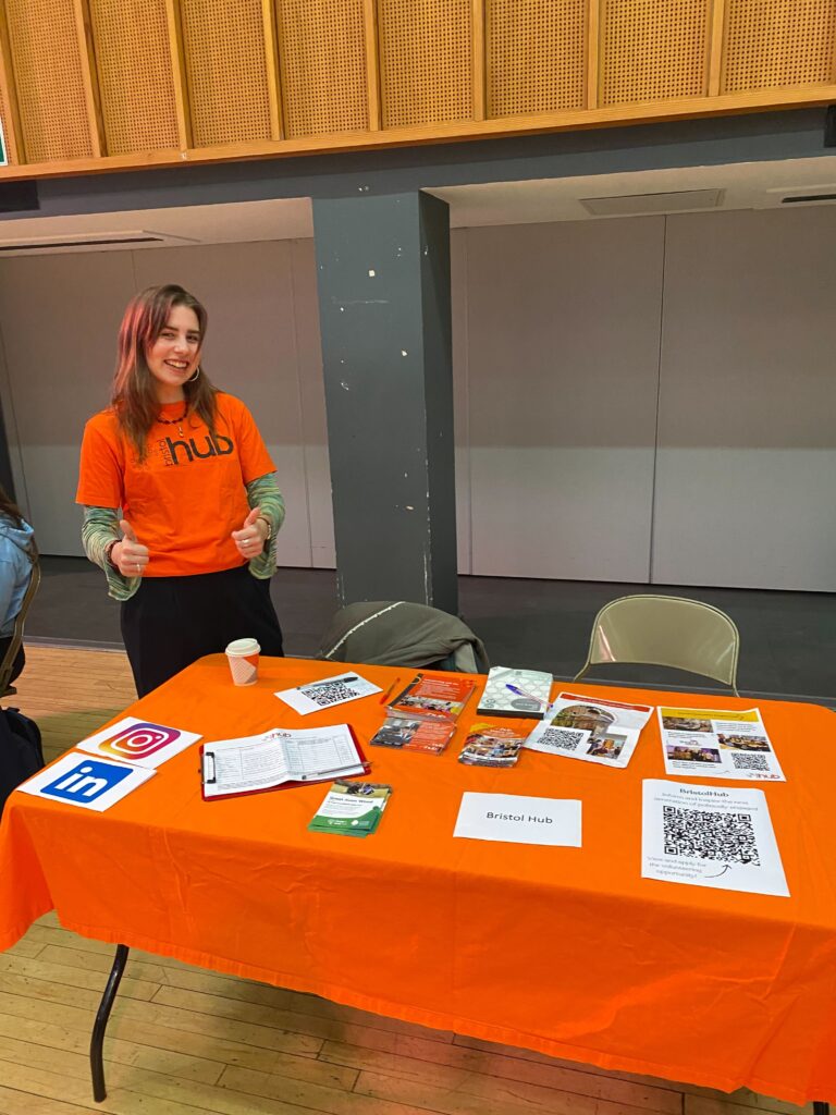 ID: a volunteer wearing an orange Bristol Hub t-shirt stands behind an orange table covered with information about Hub activities.