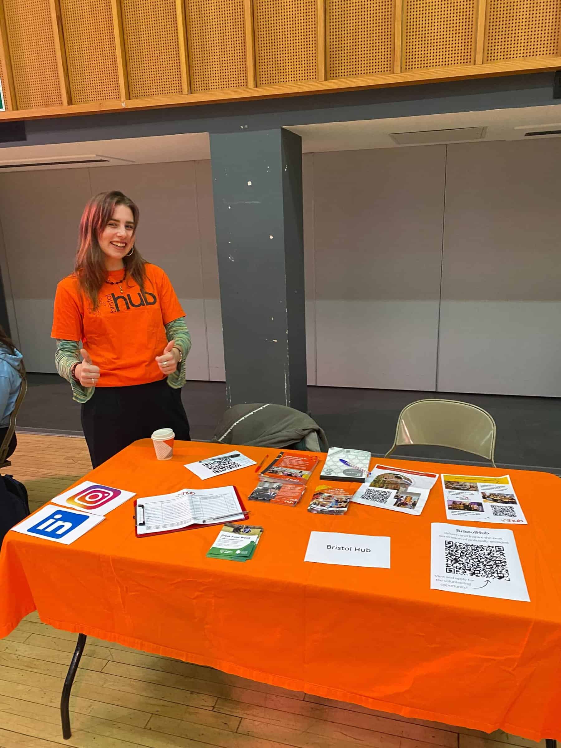 ID: a volunteer wearing an orange Bristol Hub t-shirt stands behind an orange table which is full of information about Bristol Hub activities.
