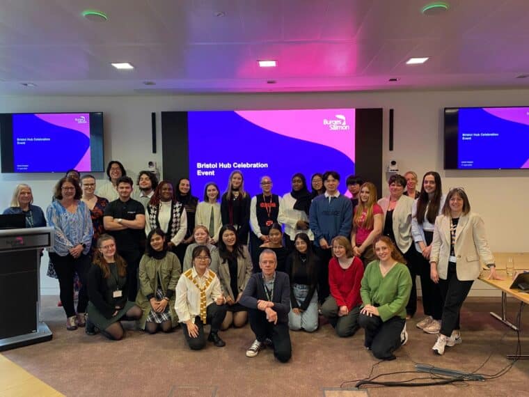 ID: SIP teams stand in front of a projector screen with text reading 'Bristol Hub Celebration Event' 